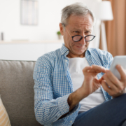Poor Eyesight. Senior Man Squinting Eyes Reading Message On Phone Wearing Eyeglasses Having Problems With Vision Sitting On Couch. Ophtalmic Issue, Bad Sight In Older Age, Macular Degeneration Concept