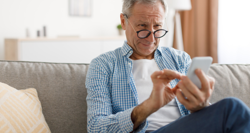 Poor Eyesight. Senior Man Squinting Eyes Reading Message On Phone Wearing Eyeglasses Having Problems With Vision Sitting On Couch. Ophtalmic Issue, Bad Sight In Older Age, Macular Degeneration Concept